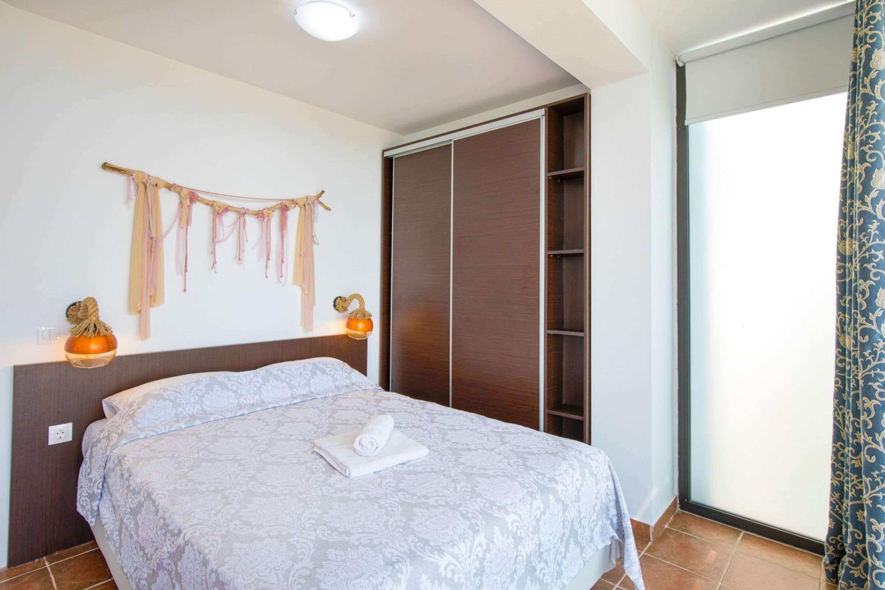 Una cama bien tendida con una colcha estampada, un armario de madera y un ventanal con cortina.