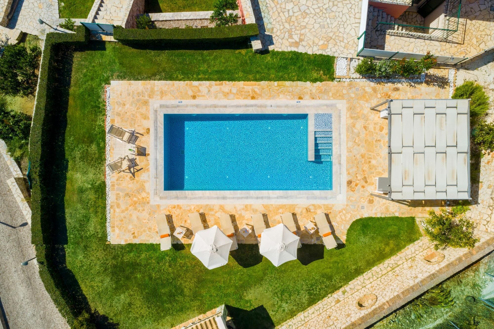 Vista aèria d'una piscina rectangular amb terrassa de pedra, envoltada d'un jardí ben cuidat i una zona de seients amb ombrel·les.