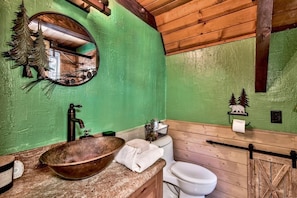 A rustic bathroom with a green wall, wooden ceiling, a round mirror, a wooden basin, a toilet, and a small shelf with a towel.