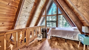 A cozy attic bedroom with a sloped wooden ceiling, a large window offering a view of trees, a bed, and a small desk with a lamp. - 3001r Reindeer A Frame Cabin 2 Bedroom Cabin by RedAwning (South Lake Tahoe)