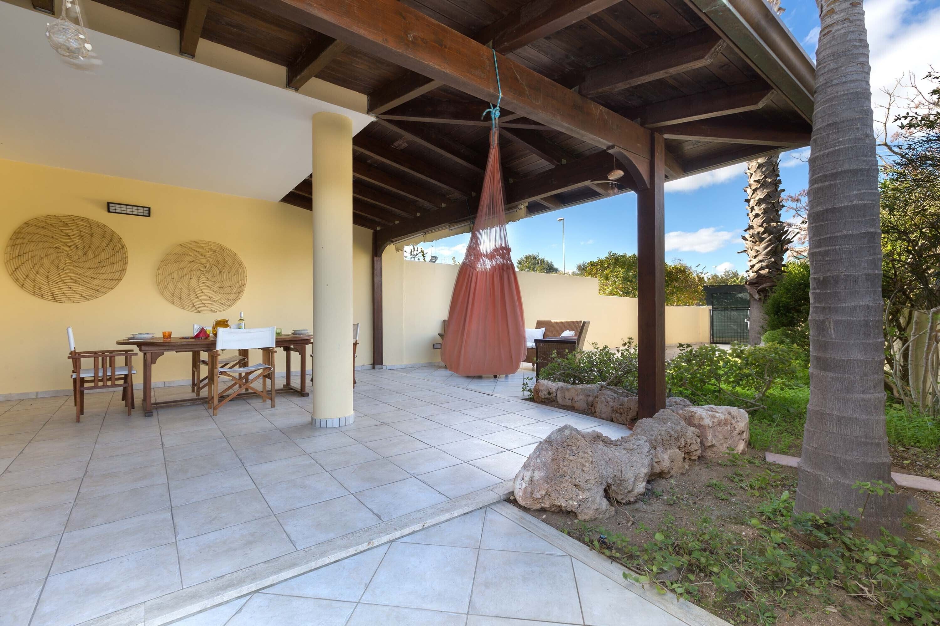 A covered patio area with a table and chairs, a hanging hammock, and a view of the surrounding landscape.