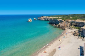 Une plage côtière aux eaux turquoise et limpides, bordée de falaises rocheuses et dotée d’un rivage sablonneux parsemé de parasols et fréquenté par des baigneurs.