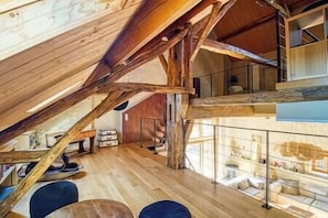 An attic room with exposed wooden beams, a loft bed, and a glass railing overlooking a lower level.