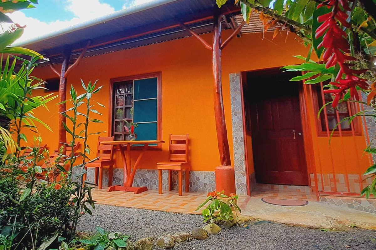 A house with an orange exterior, a wooden porch with two chairs, a window, and a door.