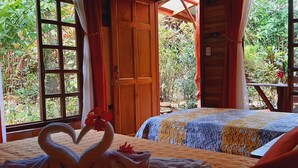 A room with a bed, a heart-shaped towel arrangement, and large windows with a view of greenery. - Jardín entre Montañas (Bijagua)