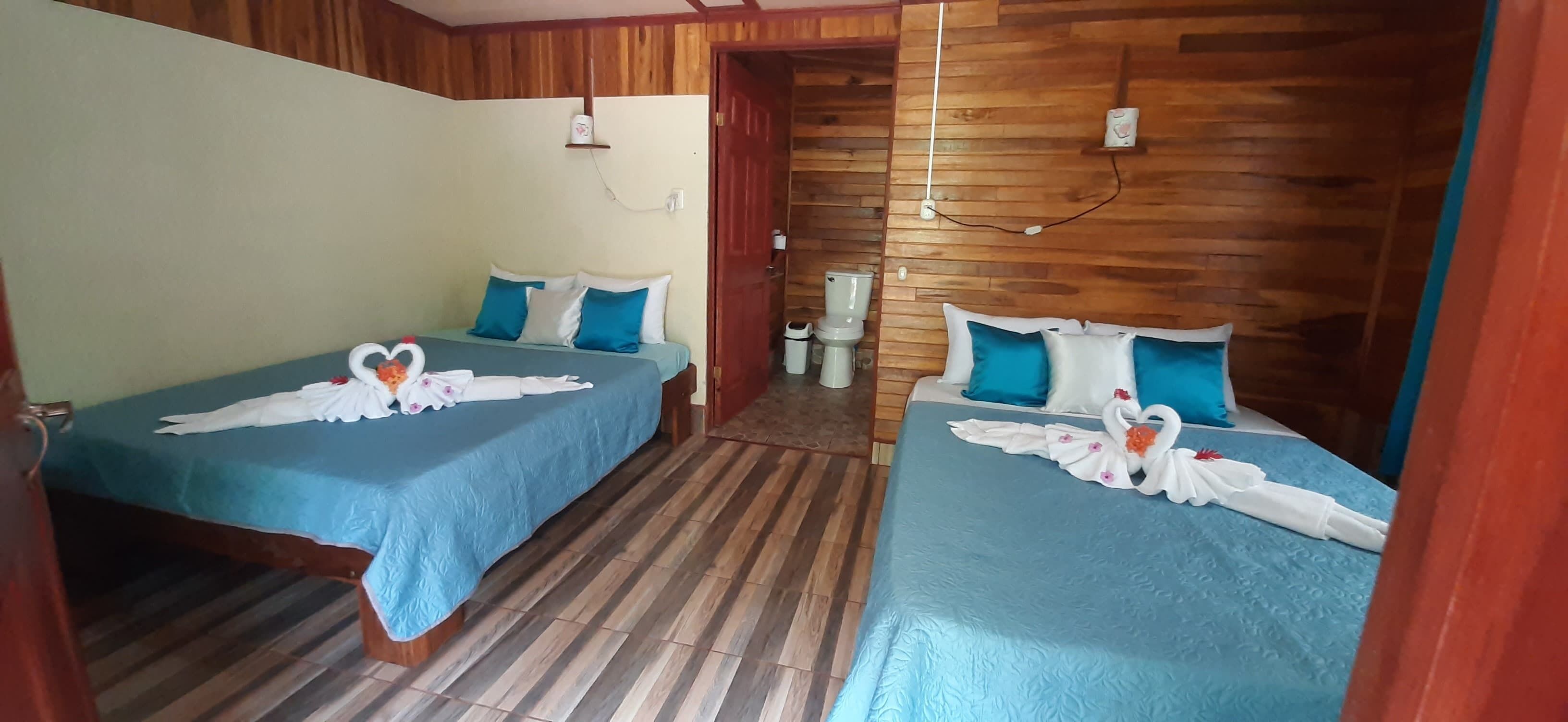 Een kamer met twee bedden, elk met een blauwe spreideken en witte kussens, en een handdoek met een zwanendesign. Er ligt een houten vloer en de wanden zijn bekleed met houten panelen.