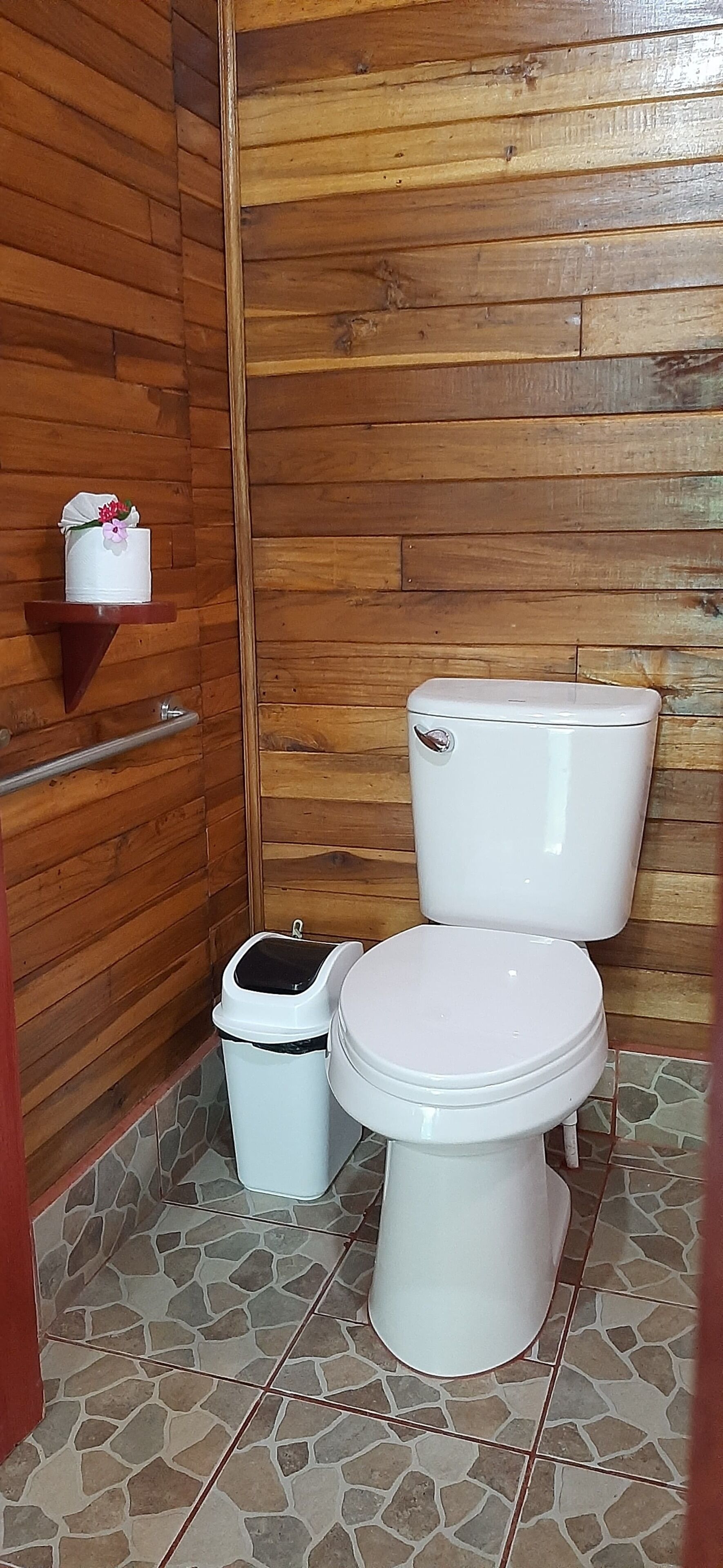 Un baño con pared de madera, un inodoro blanco, un cubo de basura y una repisa con un objeto decorativo.