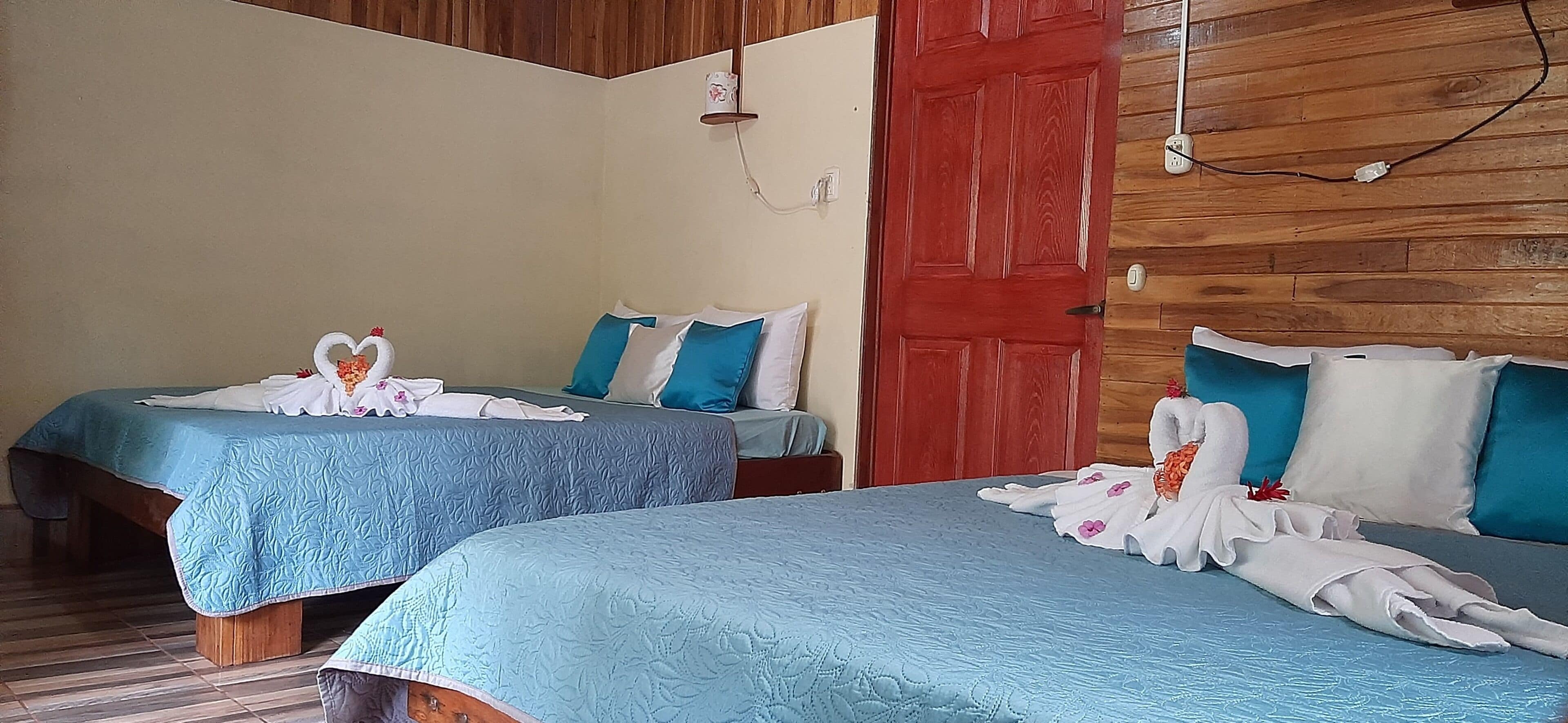 Two single beds with blue bedding and heart-shaped towels, a red door, and wooden paneling on the walls.