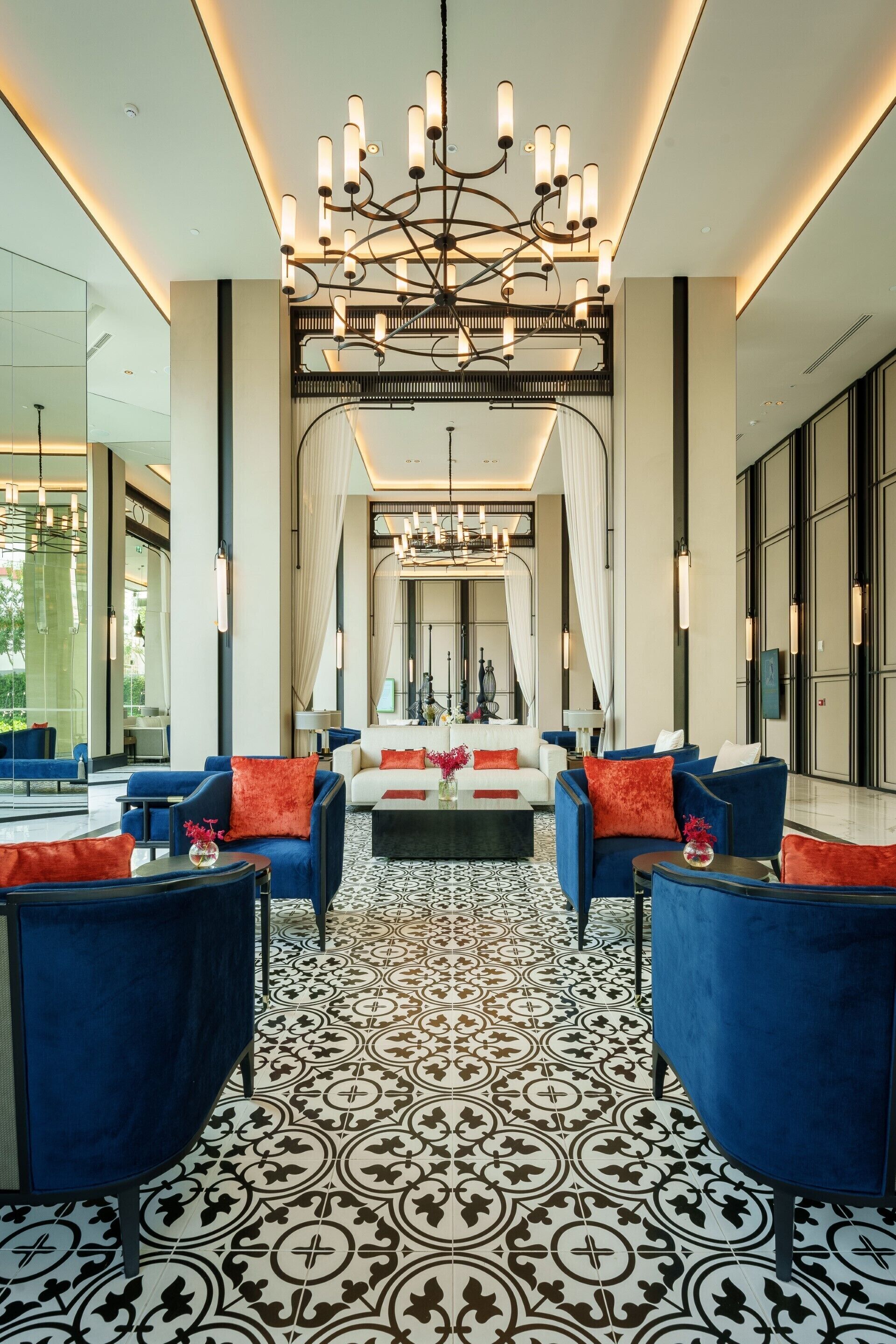 A modern lobby with a patterned floor, blue sofas, a central chandelier, and a large mirror.
