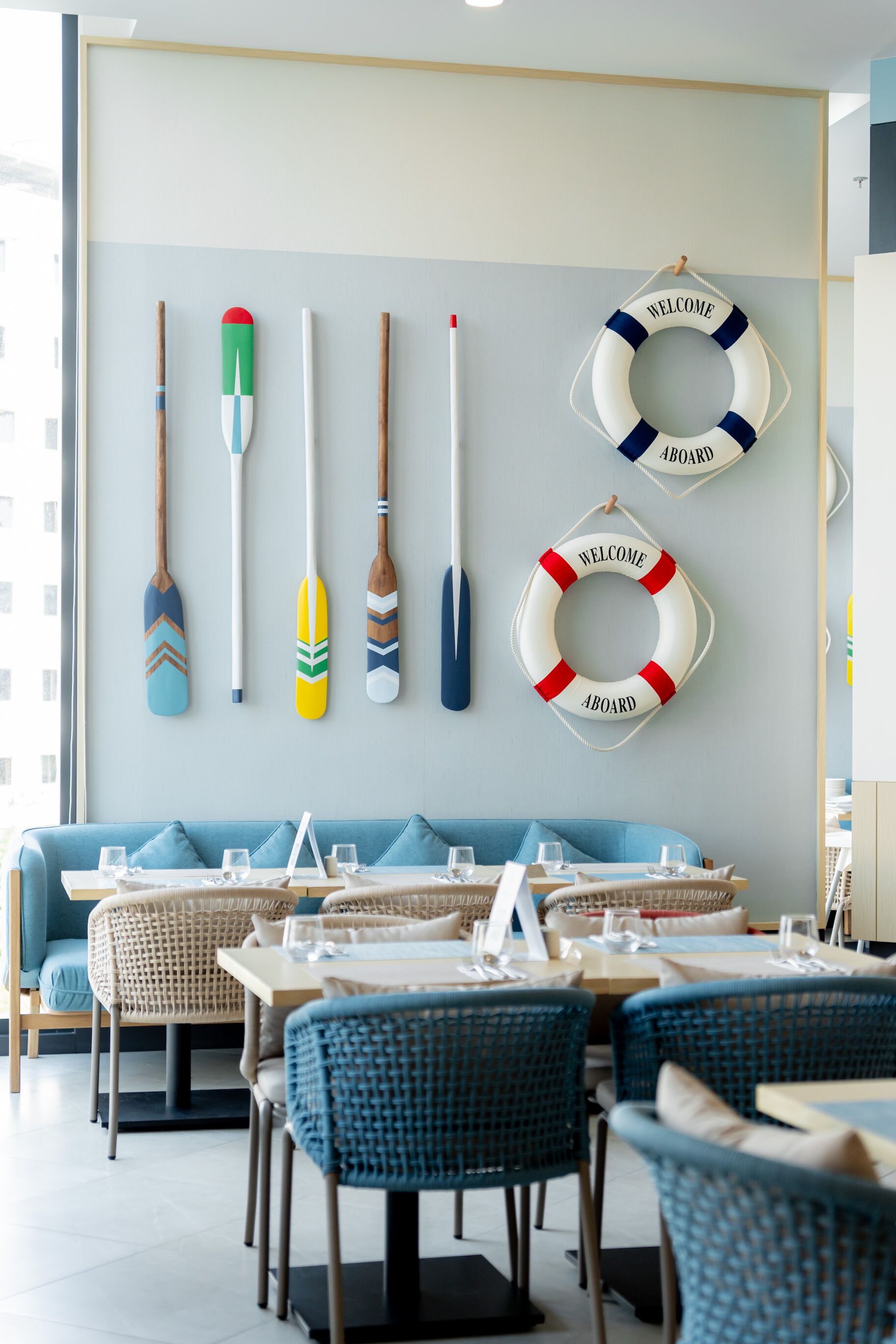 Uma sala de jantar com um sofá azul, cadeiras de vime e mesas preparadas para refeições.