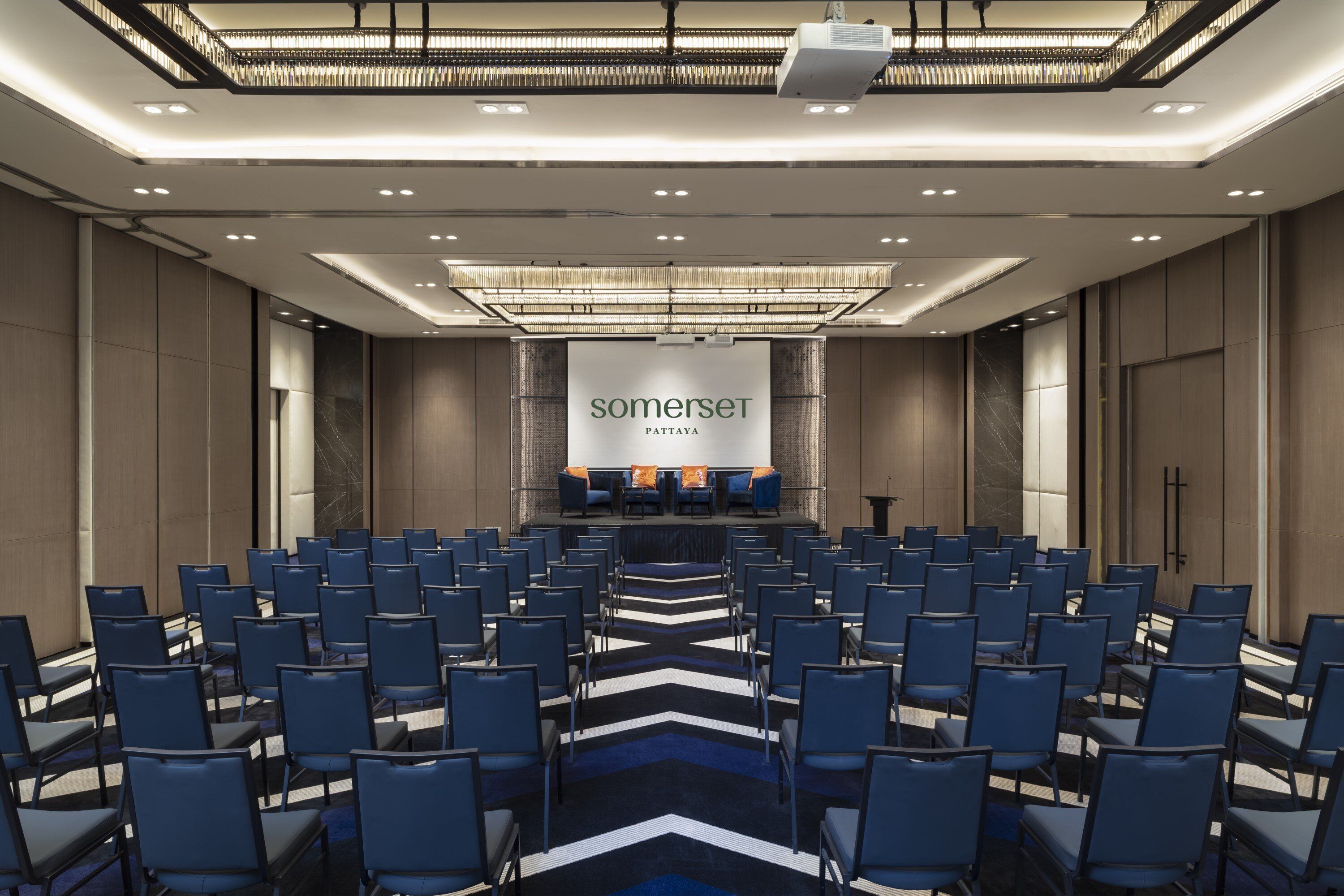 A conference room with rows of chairs, a stage with a screen displaying 'somerset pttaya', and a modern ceiling design.