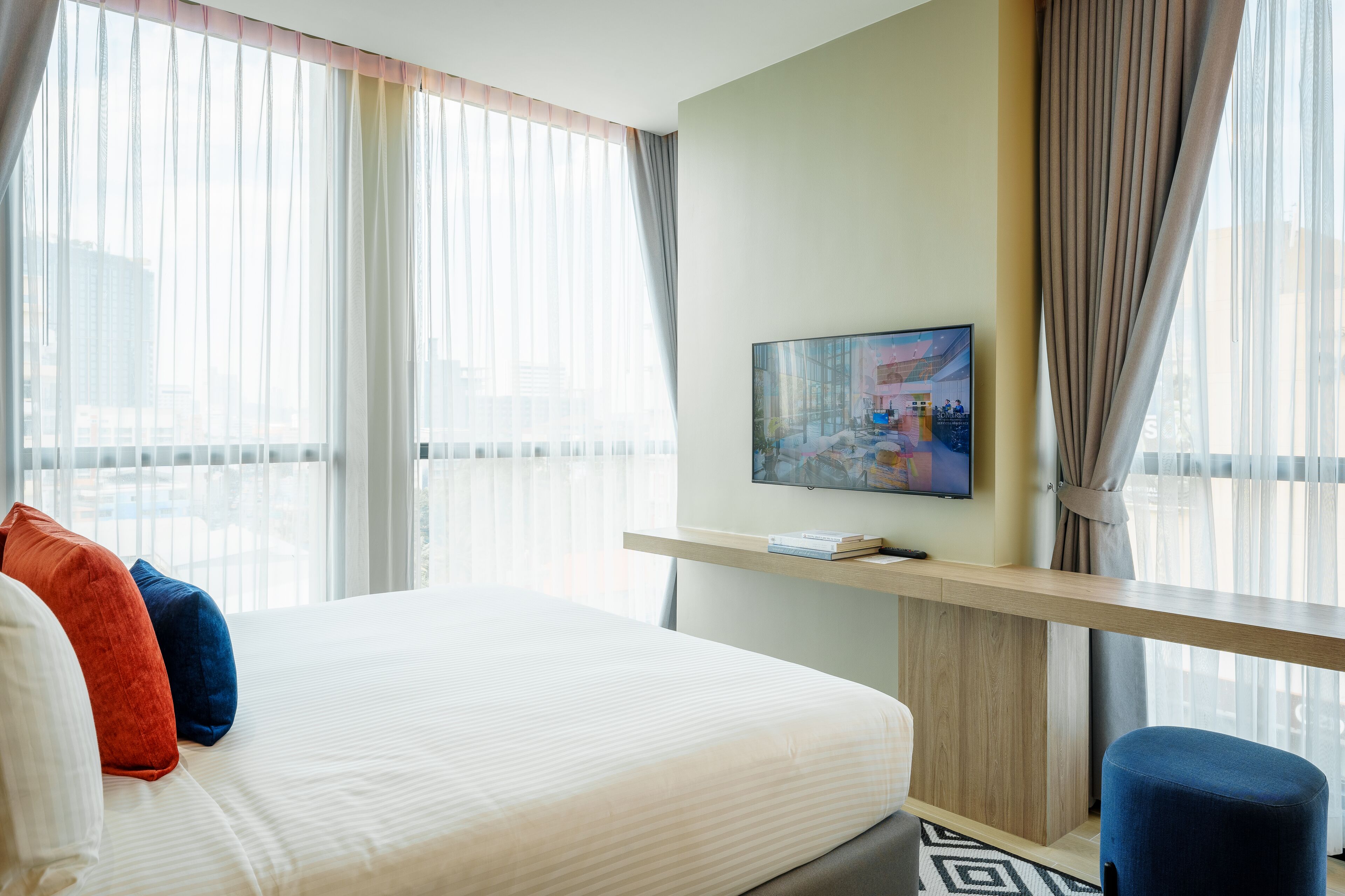 A hotel room with a large bed, a TV on a wooden shelf, and a city view through sheer curtains.