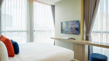 A hotel room with a large bed, a TV on a wooden shelf, and a city view through sheer curtains.