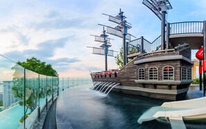 Una fuente con forma de barco y una cascada, rodeada de barandillas de vidrio y vista del paisaje urbano.