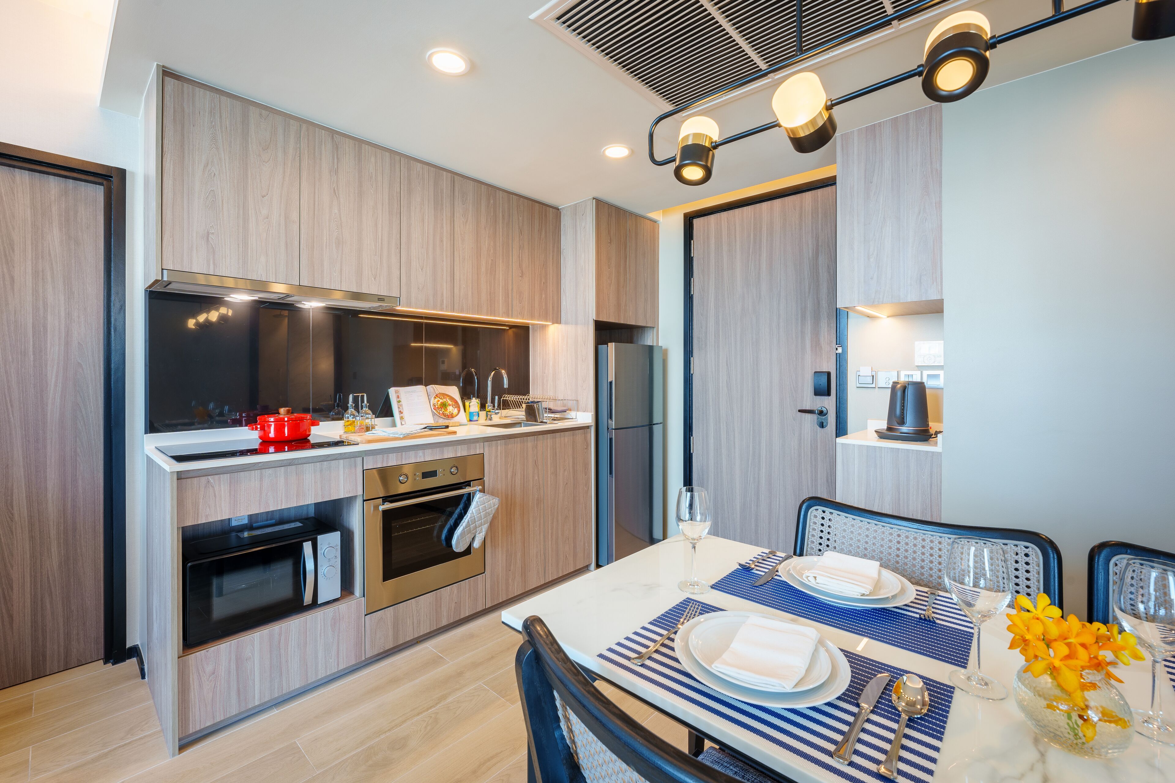 Una cocina moderna con electrodomésticos integrados, un comedor con mesa dispuesta para dos personas y un pequeño rincón de estar con una silla.