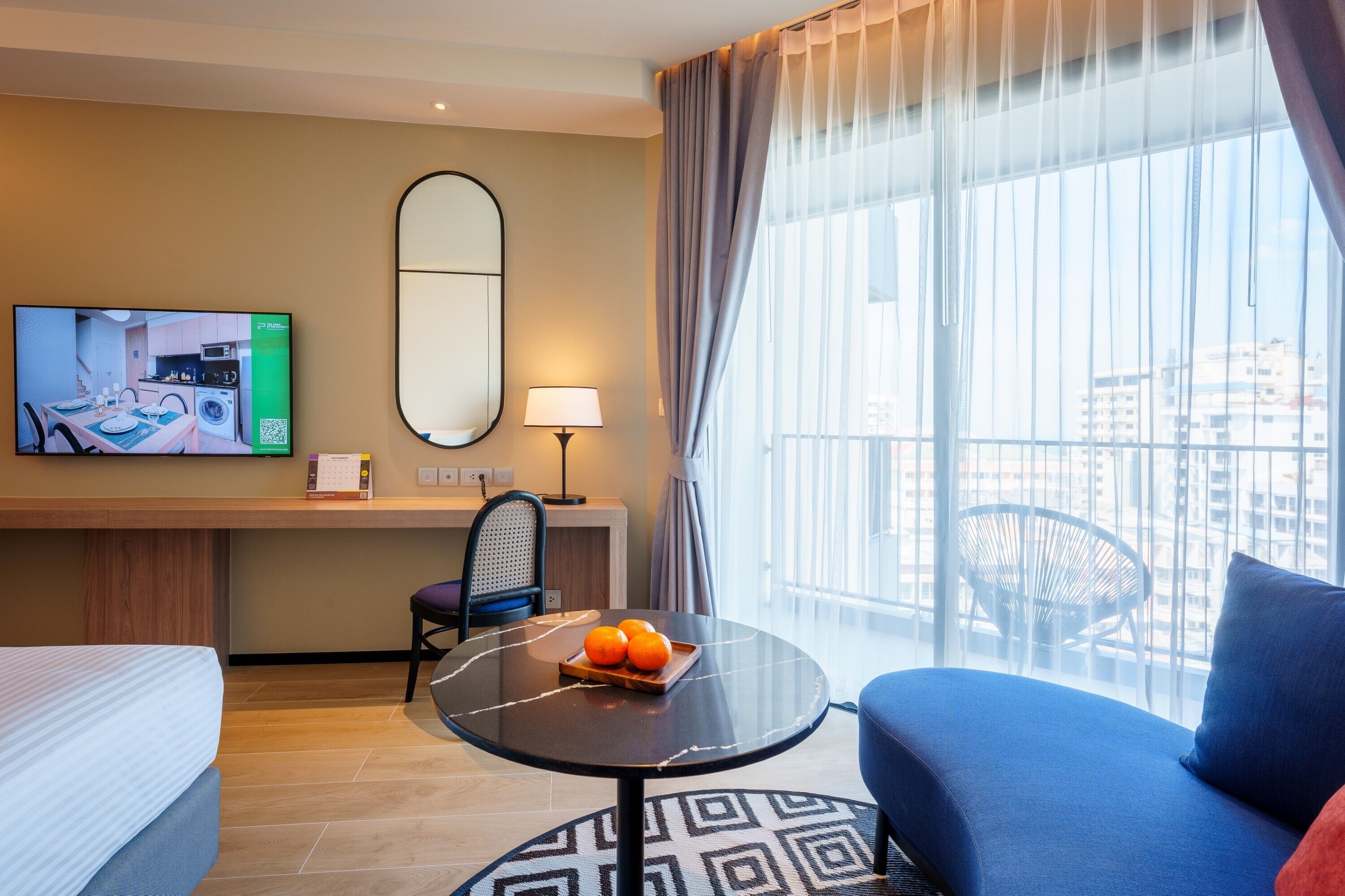 A hotel room with a bed, a sofa, a small table with oranges, a TV, a mirror, a chair, and a balcony with a view of buildings.