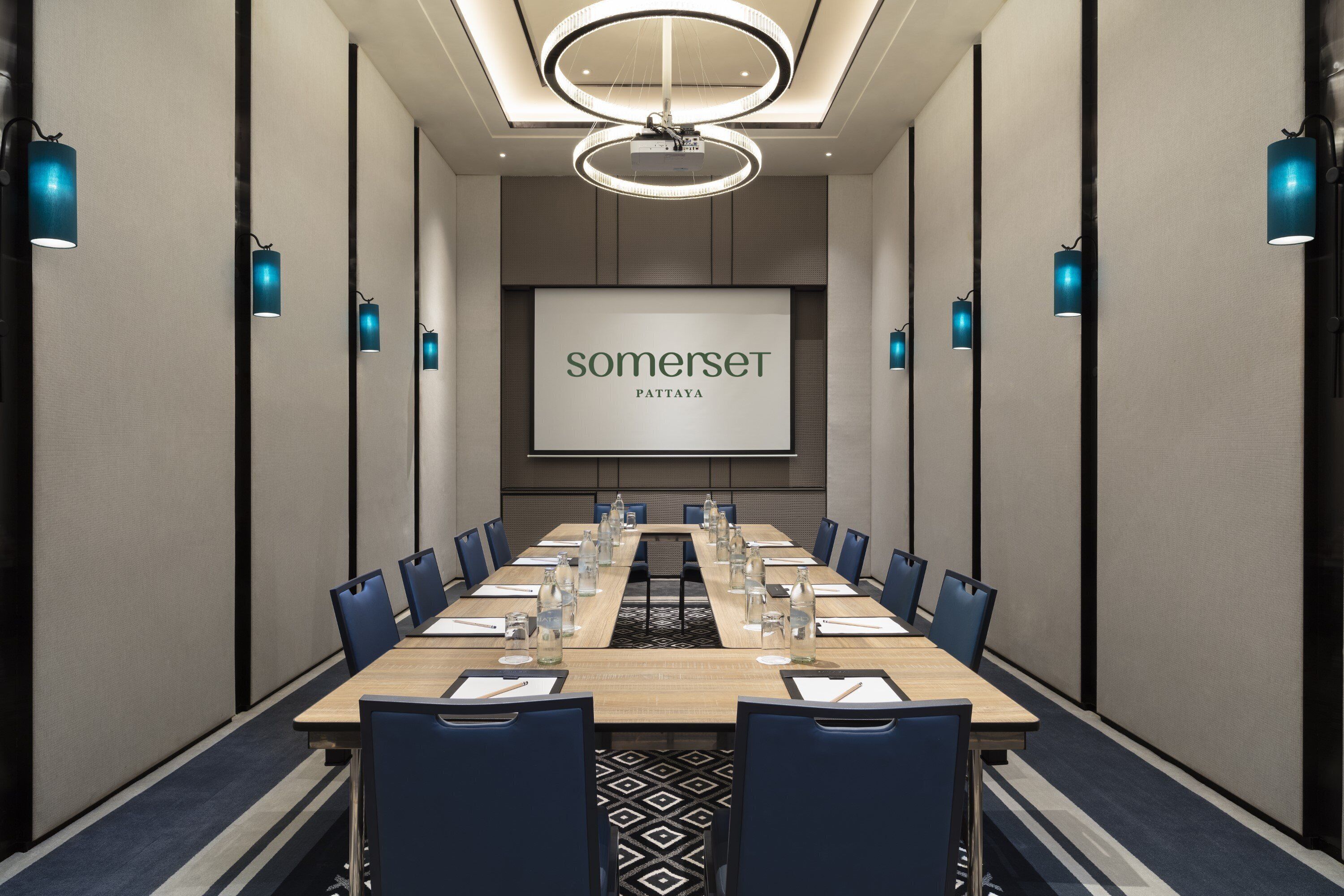 A conference room with a long table, blue chairs, and a screen displaying "somerset pattiya".