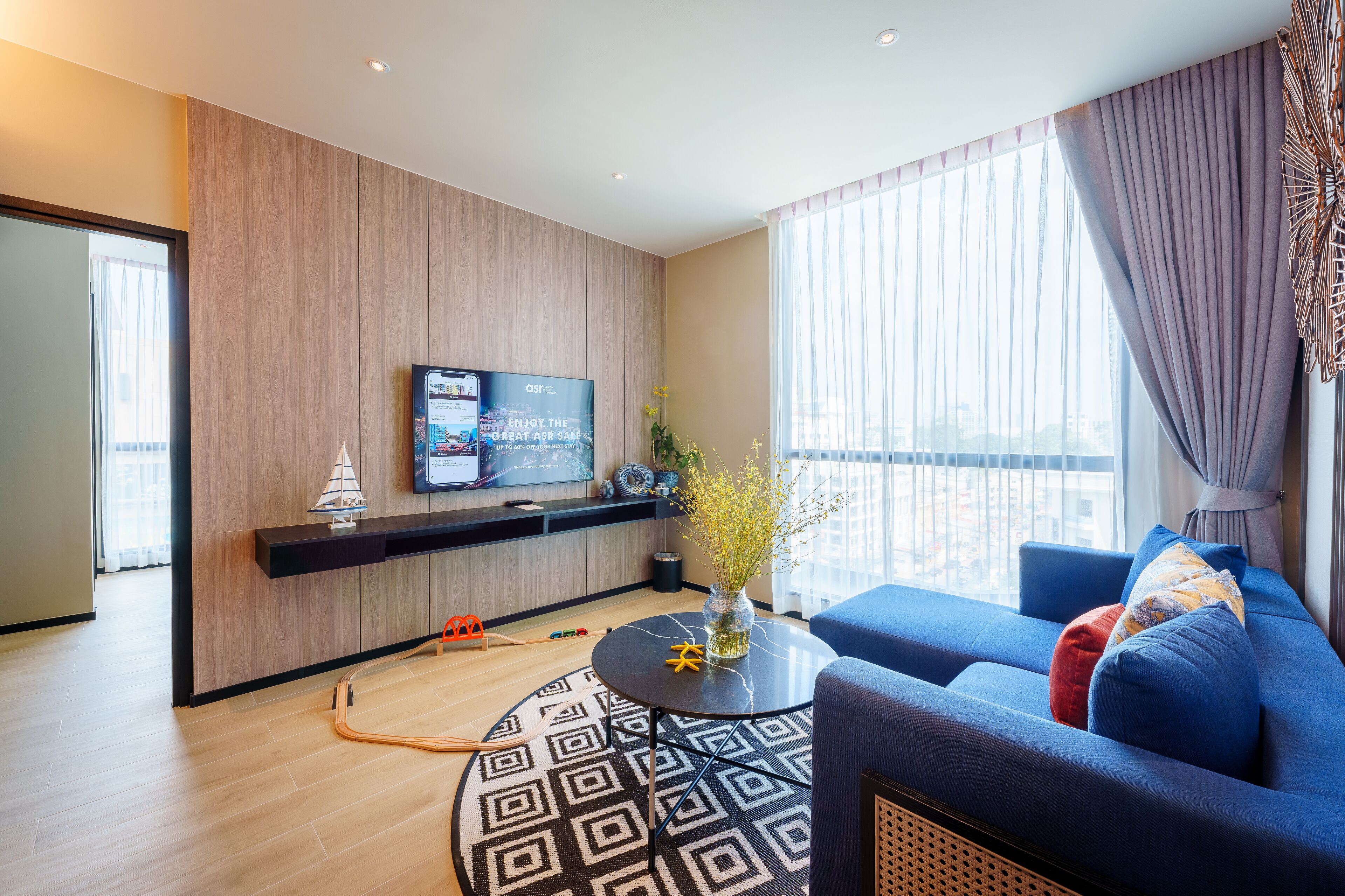 Un saló modern amb televisió de pantalla plana, un sofà blau, una taula de centre rodona i una finestra gran amb cortines.