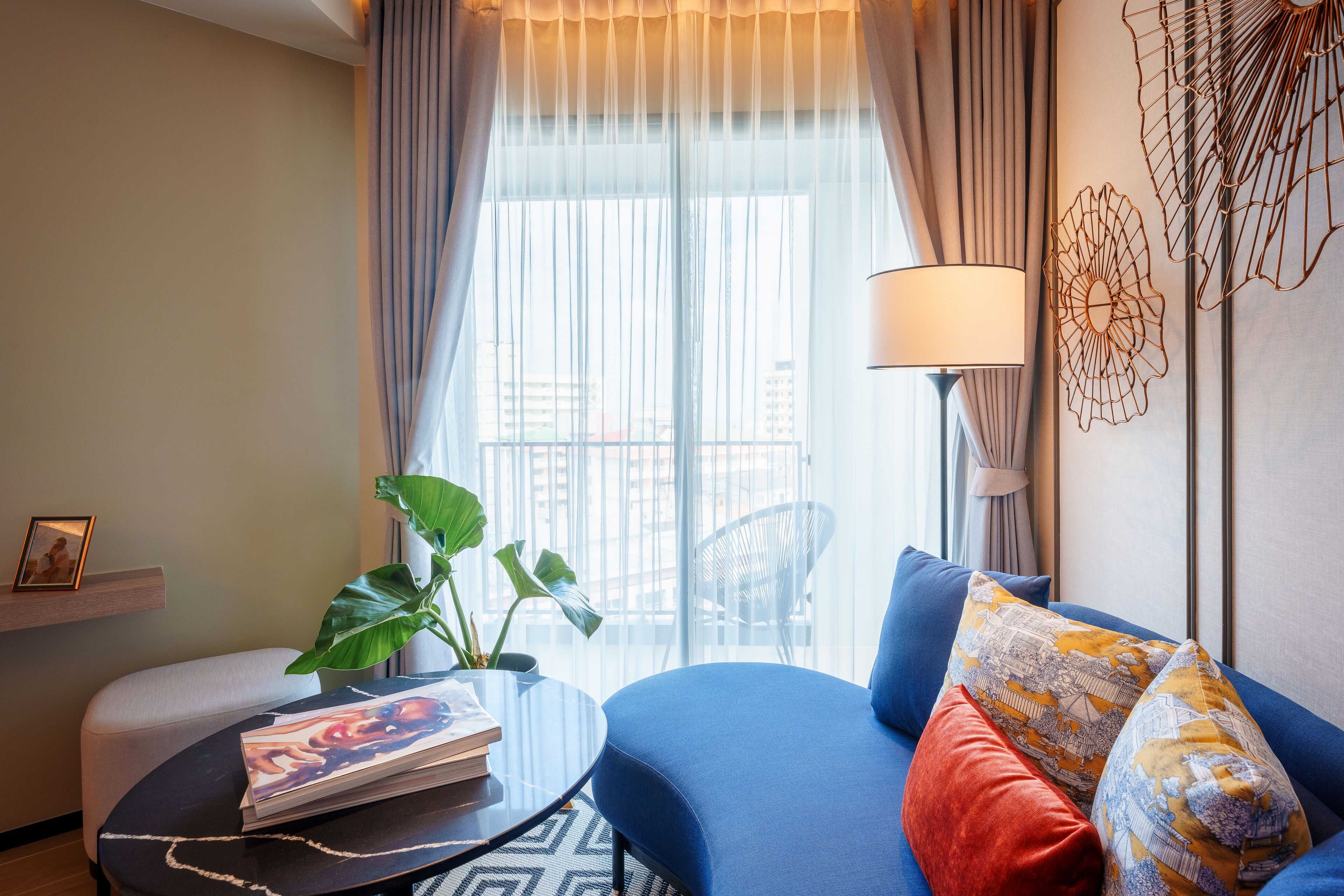 A modern living room with a blue sofa, a round coffee table, a lamp, a plant, and a view of the city through large windows.