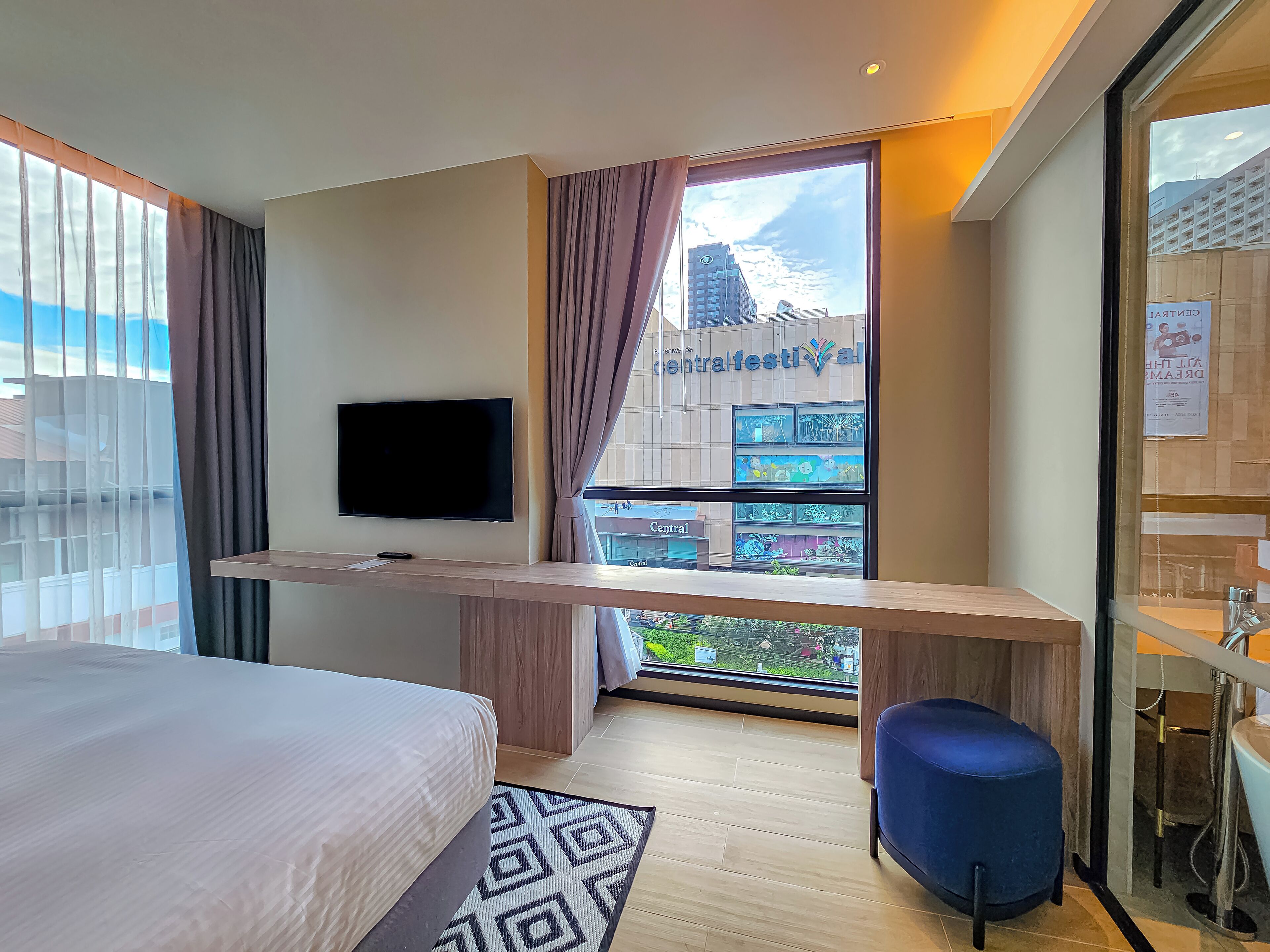 A hotel room with a bed, a TV mounted on the wall, a desk, a blue chair, and a bathroom visible through a glass door.