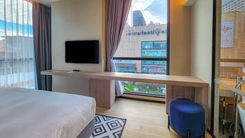 A hotel room with a bed, a TV mounted on the wall, a desk, a blue chair, and a bathroom visible through a glass door.