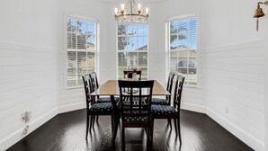 A dining room with a wooden table and chairs, large windows, and a chandelier.