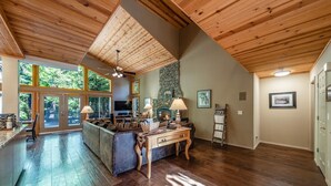 Een ruime woonkamer met houten plafond, een stenen openhaard en een groot raam met uitzicht op groen.