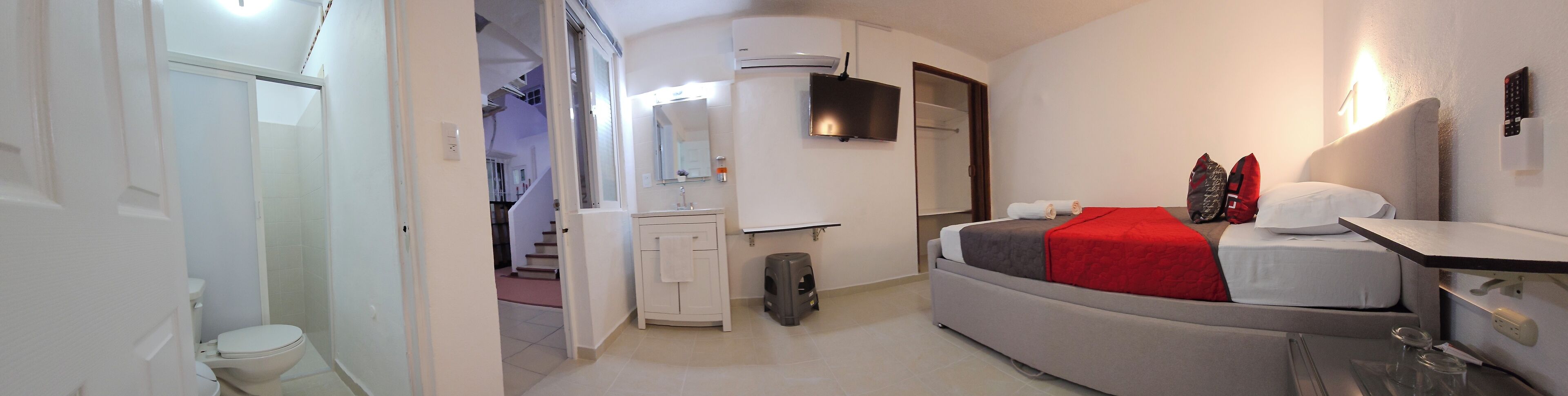 A compact bedroom with a curved bed, a bedside table, a mirror, a TV, and a small stool.