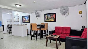 Uma sala de estar com um sofá vermelho, um sofá preto, uma mesa de centro de madeira e uma área de jantar com mesa e cadeiras brancas.