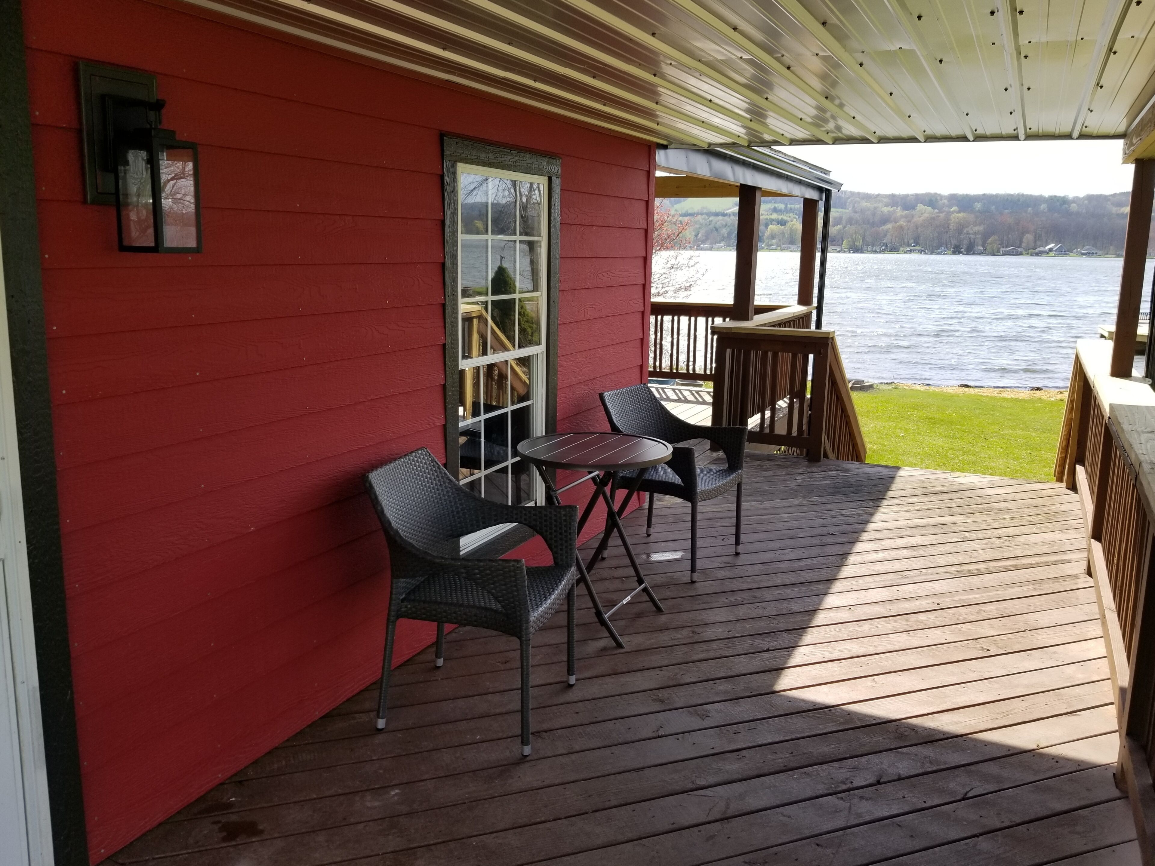 Un bâtiment rouge doté d’une terrasse en bois, de deux fauteuils en osier, d’une petite table et offrant une vue sur un lac.