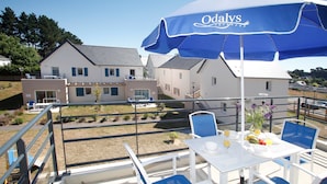 A balcony with a table set for two, a blue umbrella, and a view of houses in the background.