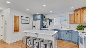A modern kitchen with a central island, bar stools, blue cabinets, and a white refrigerator. - The Gathering Place for Paws, Friends, and Family (Orem)
