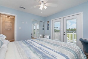Un dormitorio con cama, ventilador de techo, puerta que da a un balcón y un cartel con motivos playeros en la pared.