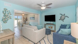 A living room with a ceiling fan, a TV, and a dining area with chairs and a table. - Seaglass Shores, Steps to the Beach (Cape Canaveral)