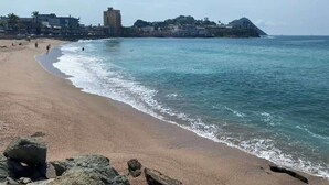 Una playa con primer plano rocoso, mar tranquilo y edificios al fondo.