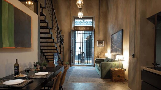 Comedor con una mesa dispuesta para dos personas, una escalera y una sala de estar con sofá y lámpara.