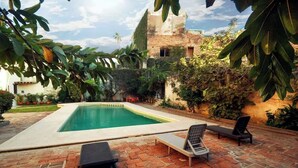 Una piscina rectangular con sillones de descanso y una mesa, rodeada de follaje y un muro de ladrillo.