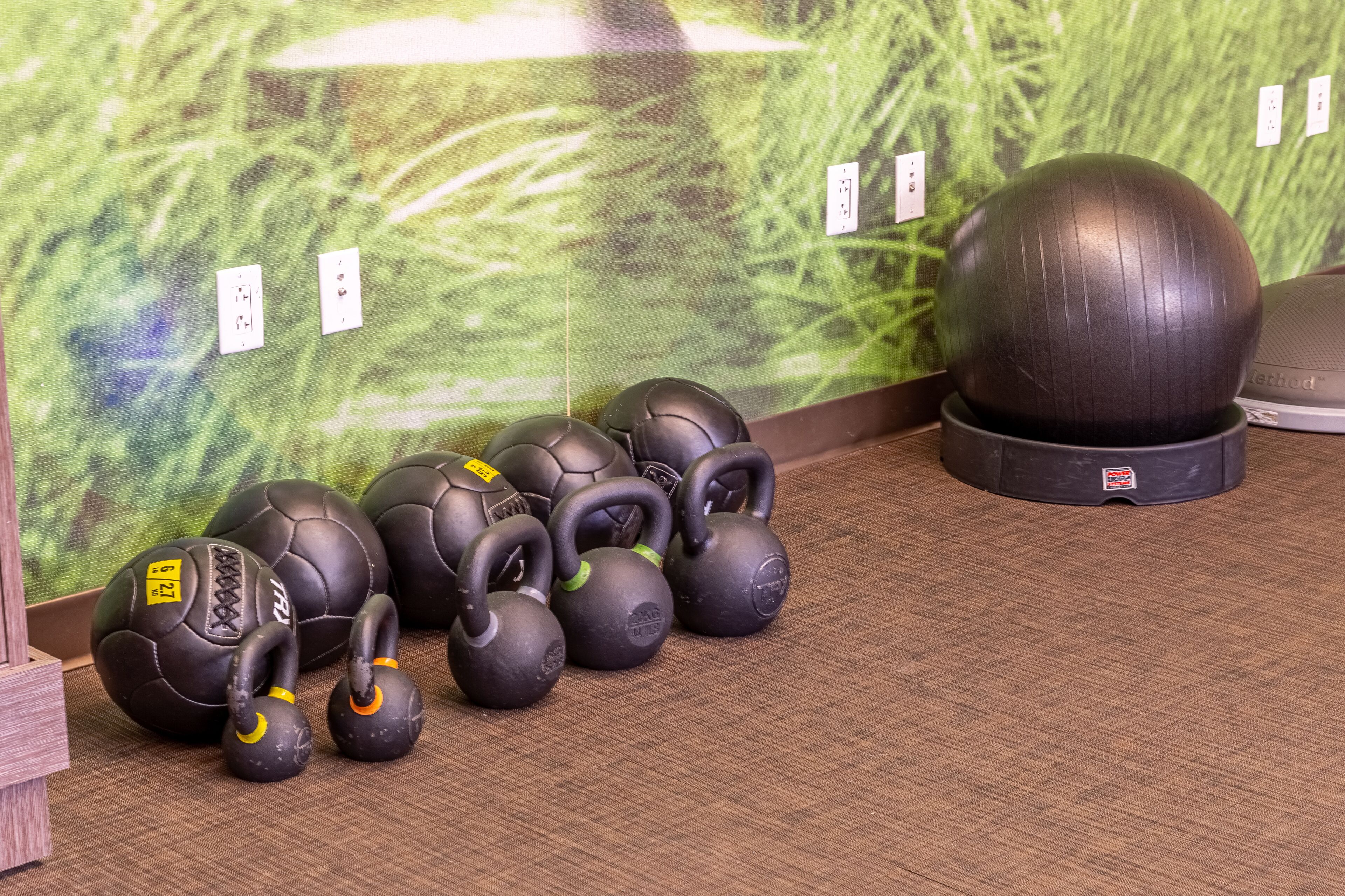 Verschiedene Kettlebells und ein Medizinball auf einem Holzboden, im Hintergrund eine Wand mit grüner Struktur.