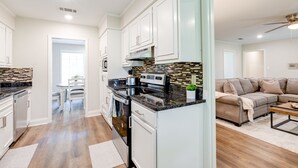 Une cuisine moderne avec des armoires blanches, un comptoir noir et un dosseret carrelé. On y trouve un coin repas avec une table et des chaises, ainsi qu’un salon avec un canapé beige et une table basse.