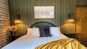Ein Bett mit weißer Steigbügeldecke und goldener Tagesdecke, begleitet von zwei Nachttischen mit gewobenen Lampen. Über dem Bett hängt eine dekorative Wandgestaltung.