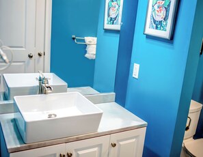 Ein Badezimmer mit zwei weißen Waschbecken, einem Spiegel und einer Toilette. An der Wand befinden sich gerahmte Kunstwerke.