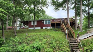 A rustic cabin with a wooden staircase leading up to a deck, surrounded by lush greenery and a small stream. - Lake of the Ozarks Secluded Vacation Home Waterfront with a Covered Dock. (Stover)