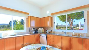 A kitchen with wooden cabinets, a dining table with a patterned tablecloth, and a view of the outdoors through large windows. - The Vines in Latchi With 3 Bedrooms and 3 Bathrooms (Neo Chorio)