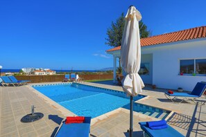 Une piscine avec des chaises longues et un parasol, une vue sur la mer et un bâtiment avec un toit en tuiles.