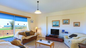 A living room with a sofa, a coffee table, and a TV. There are pictures on the wall and a view of the outdoors. - The Vines in Latchi With 3 Bedrooms and 3 Bathrooms (Neo Chorio)