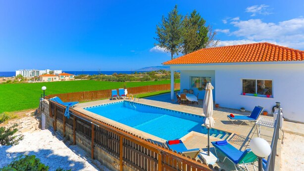 A swimming pool with lounge chairs and umbrellas, surrounded by a wooden fence and a view of the sea. - The Vines in Latchi With 3 Bedrooms and 3 Bathrooms (Neo Chorio)