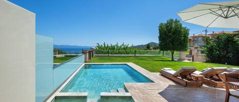 A swimming pool with a view of the sea, surrounded by a patio with lounge chairs and an umbrella.