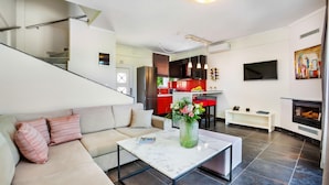A modern living room with a sofa, a coffee table, and a fireplace. There is a kitchen in the background with red cabinets and a dining area. - Sunny Villa Grande in Kassandra (Kassandra)