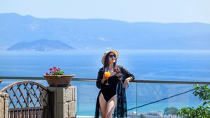 A person in a black swimsuit and a patterned robe standing by a railing with a view of the sea and distant mountains. - Sunny Villa Grande in Kassandra (Kassandra)