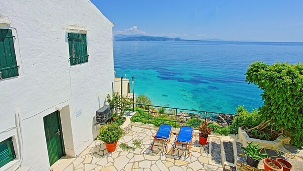 A white building with a stone patio, two blue lounge chairs, and potted plants. - Alexandra Studio in Katavolos (Corfu)